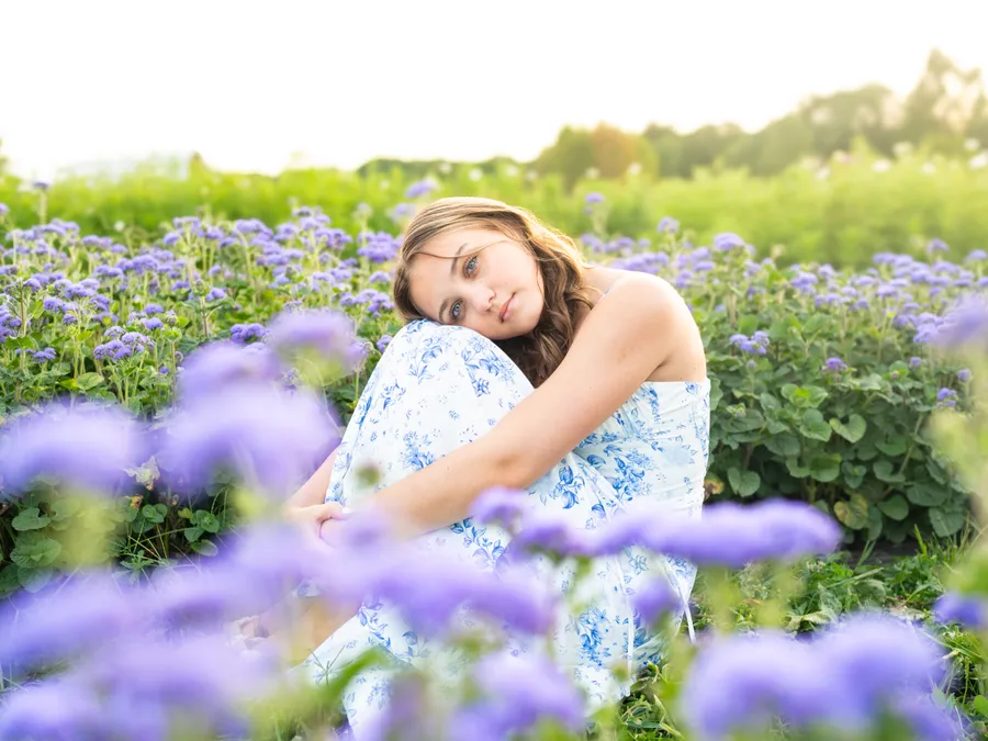 A high school senior in a white dress sitting in a sunlit field of wildflowers.